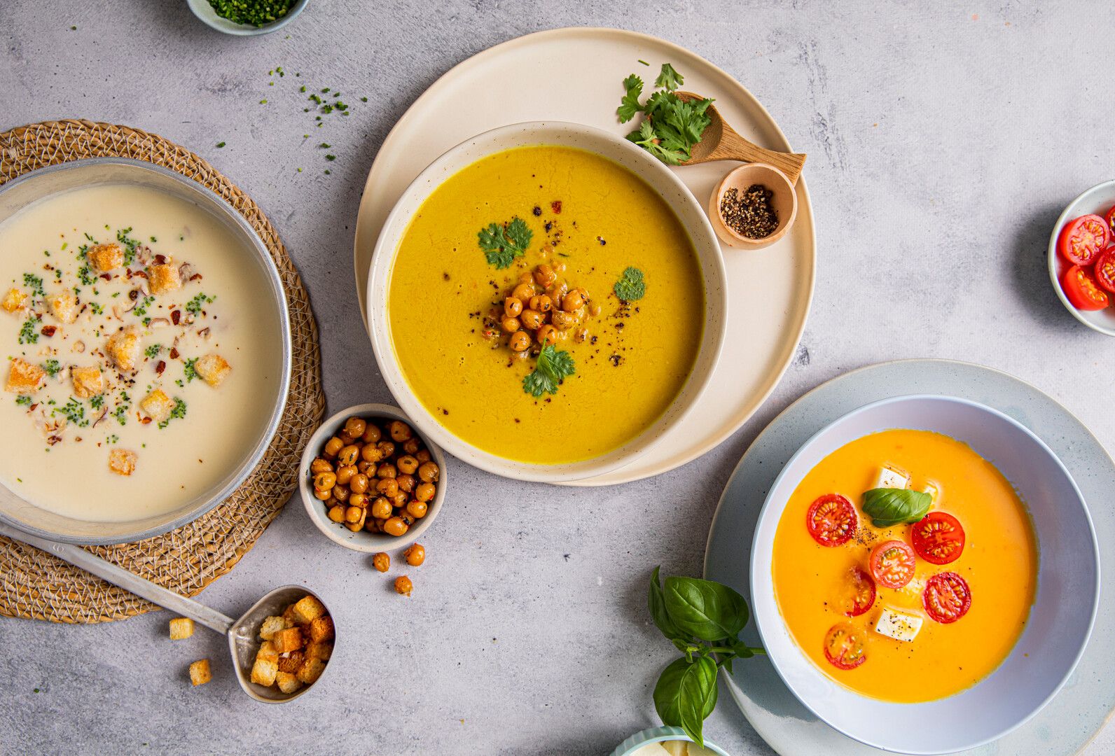 "Tre ulike kremsopper arrangert på grå bakgrunn: hvit soppssuppe, gul gulrotssuppe og oransje tomatsuppe, alle garnert med friske urter og toppinger"