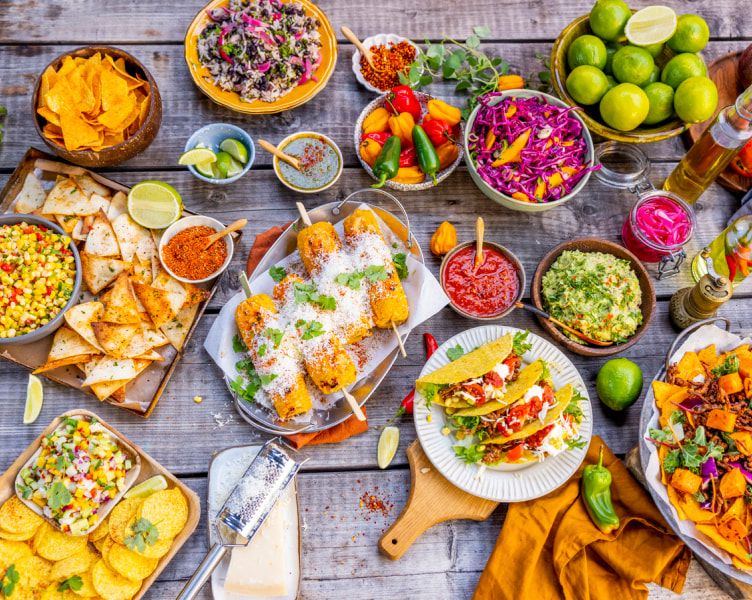Fargerikt texmex-bord med tacos, grillede maiskolber, tortillachips, guacamole, salsa, salater, lime og ulike grønnsaker. Rettene er servert på et rustikt trebord med mange små tilbehørsskåler.