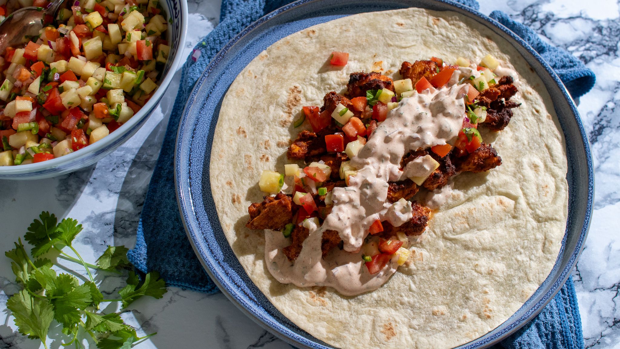 Bord med blå duk, tallerken med kyllingtaco-wraps, skål med grønnsaker og koriander.
