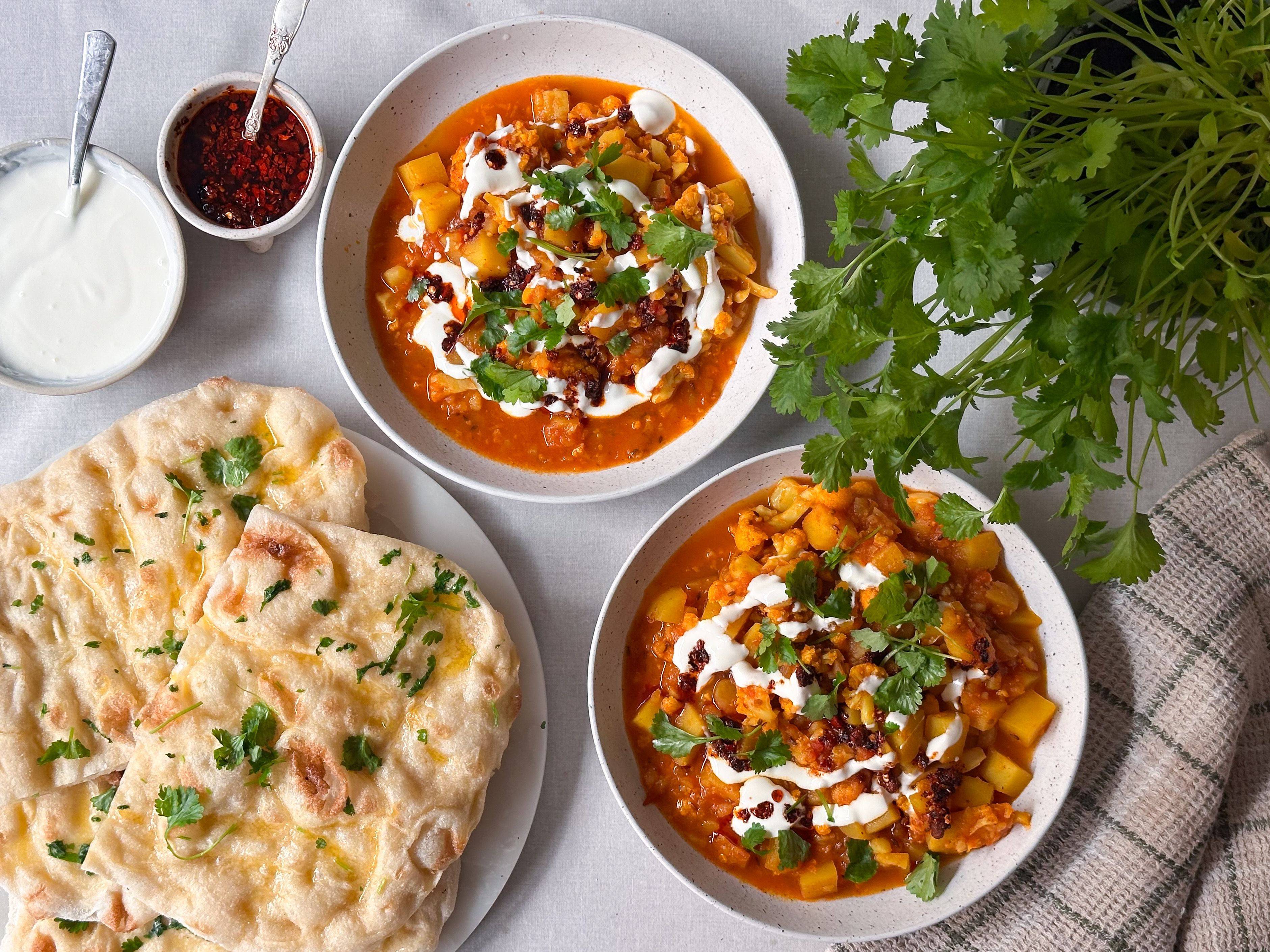 To skåler med krydret grønnsaksgryte, yoghurt og koriander, servert med naanbrød.