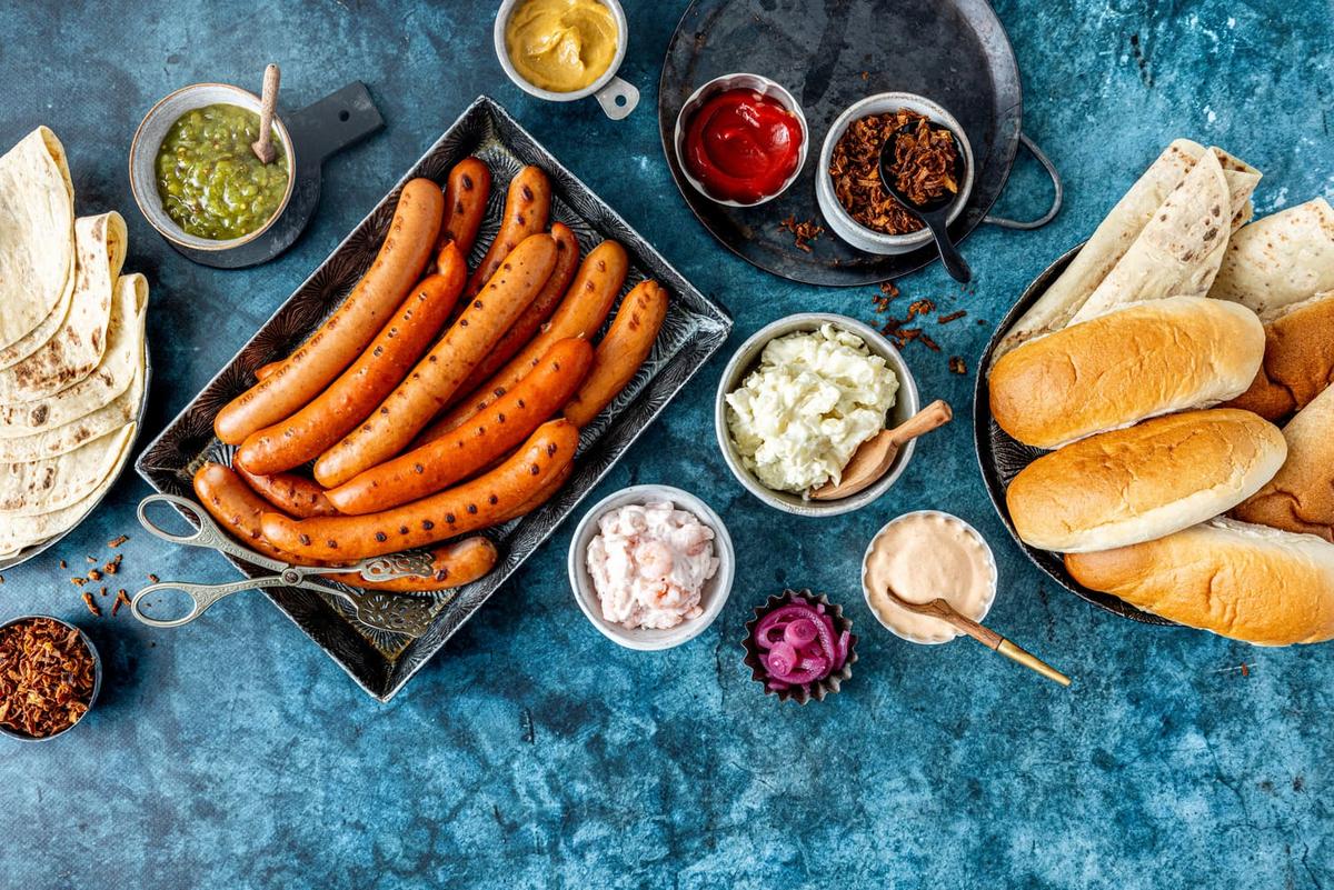 En blå bordplate dekket med smakfulle grilla pølser med tilbehør som pølsebrød, lomper og diverse av årets nye dipper.