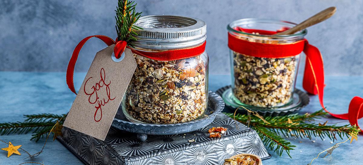 Glasskrukker fylt med müsli, frø og tørket frukt, med treskje og dryss av ingredienser på bordet.