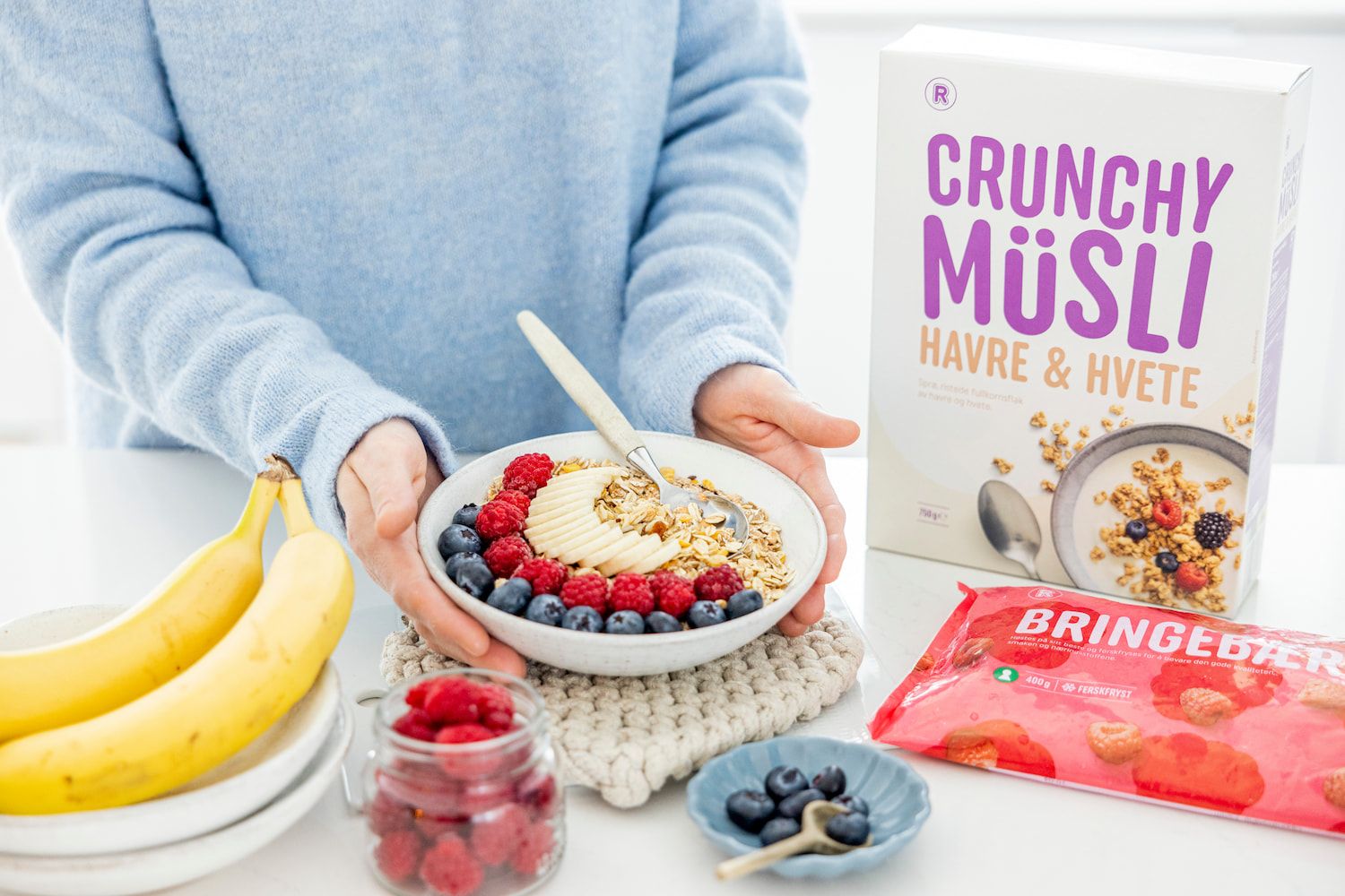 Skål med crunchy müsli toppet med banan, bringebær og blåbær holdes på et bord. Ved siden av står en pakke Crunchy Müsli Havre & Hvete, frosne bringebær, bananer og friske bær.