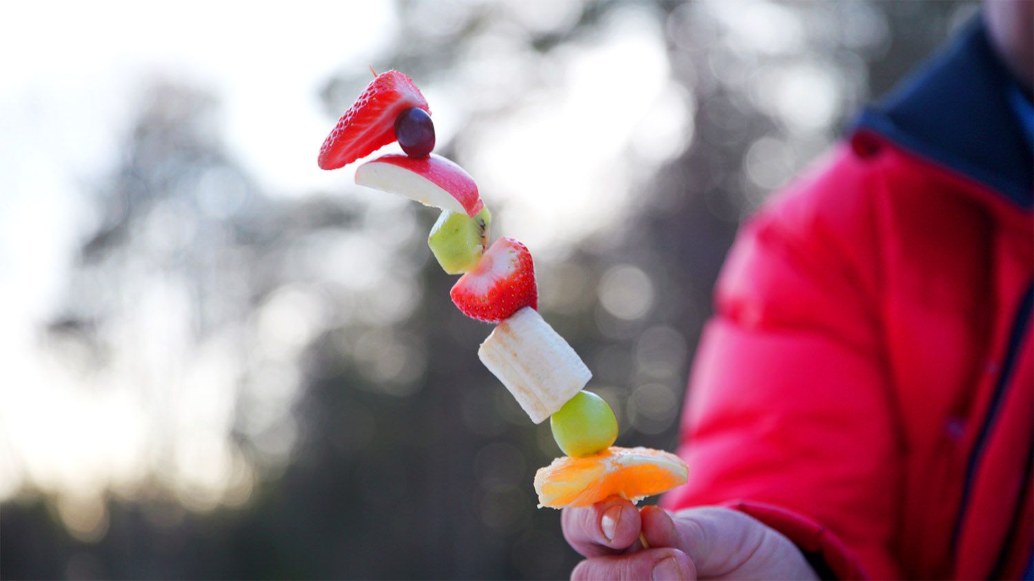 Hånd som holder et fruktspyd med jordbær, banan, druer og melon ute i vinterlys