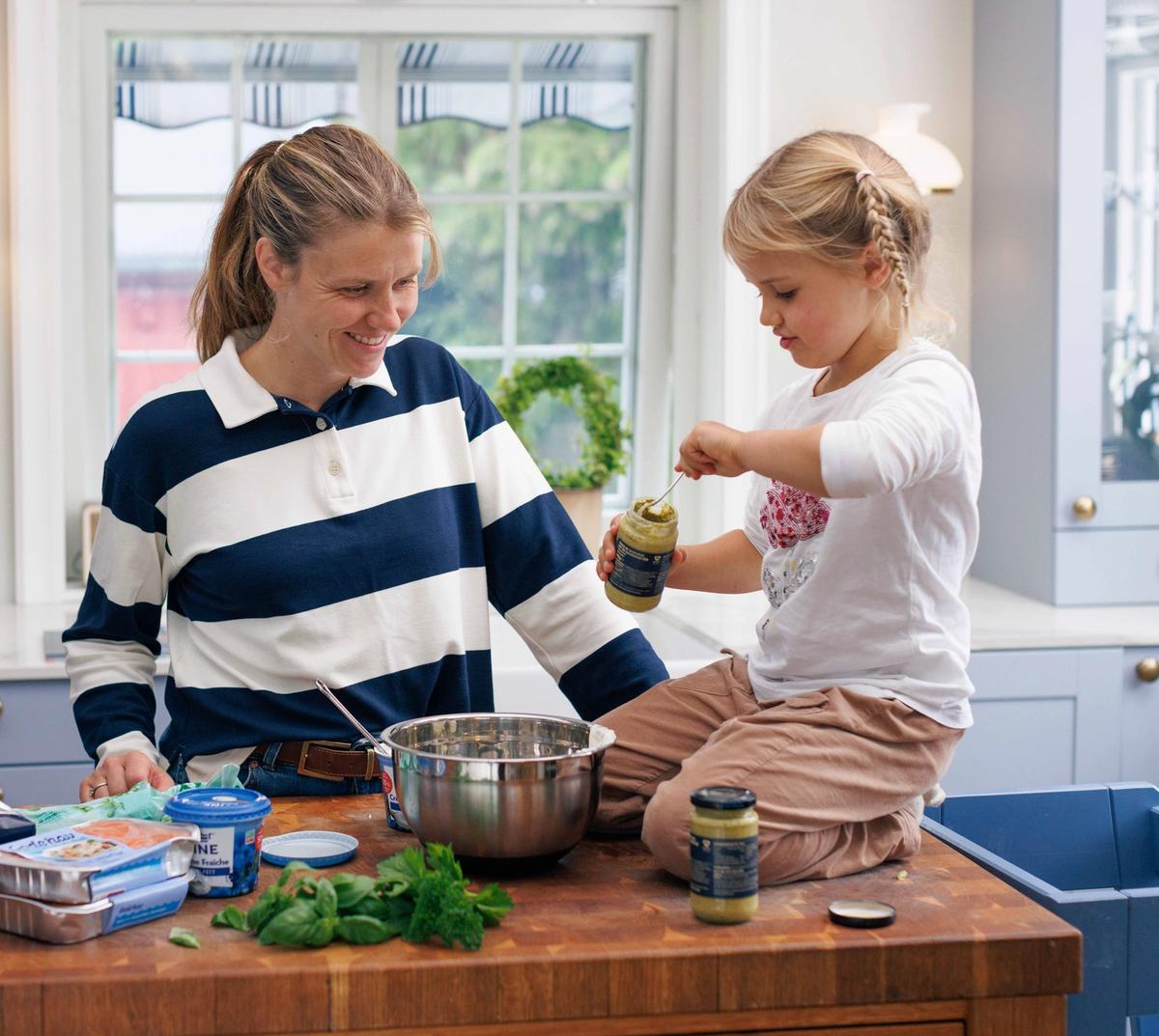 Tiril smiler til datteren, som øser pesto fra en glasskrukke ned i en bolle på kjøkkenbenken.