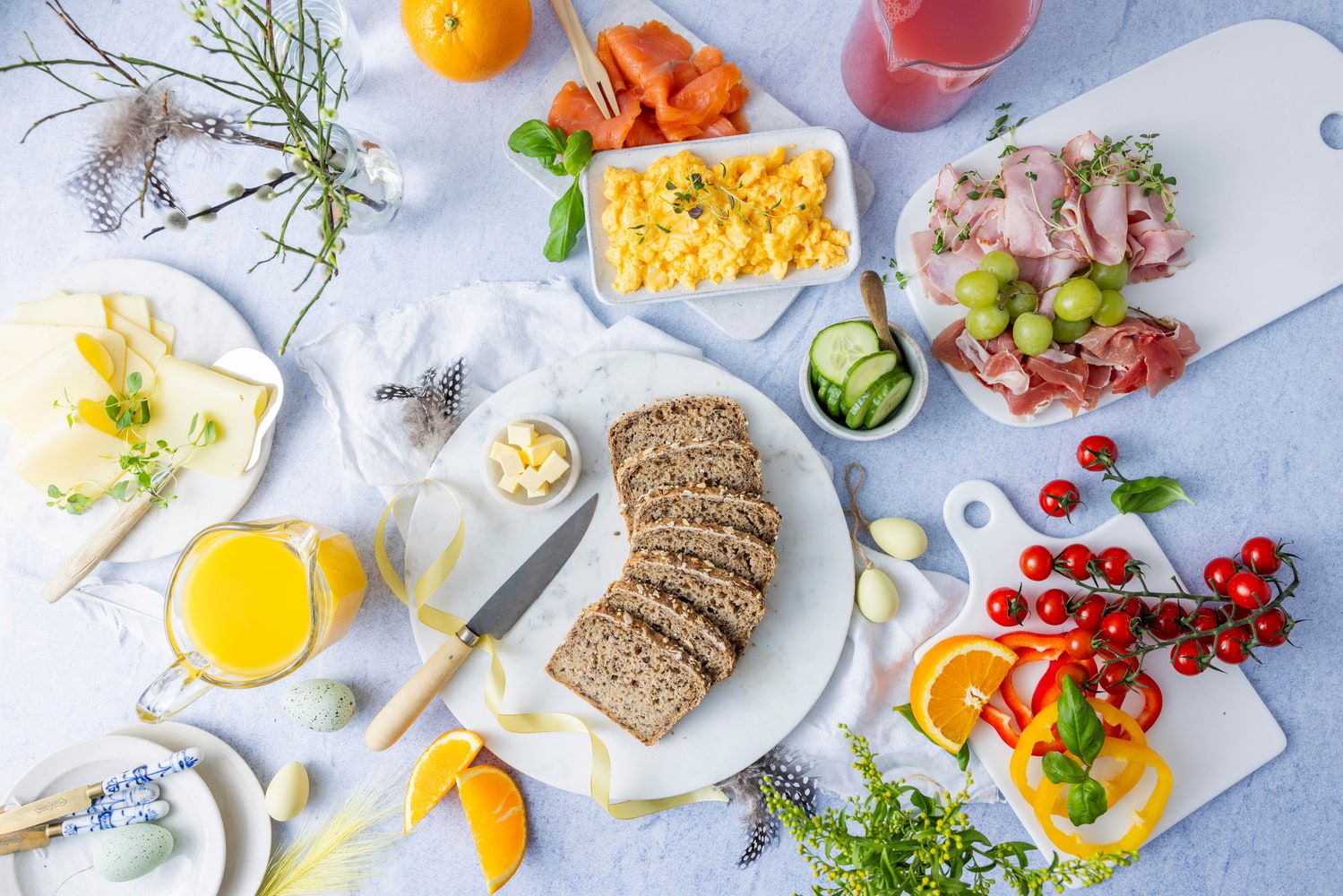 Påskefrokost med grovt brød, eggerøre, røkelaks, spekemat, ost, grønnsaker og appelsinjuice servert på et lyst bord dekorert med påskeegg og fjær.