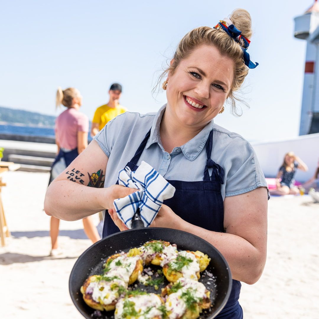 Reneé Fagerhøi holder fram en panne med smasha poteter toppet med rømmedressing, urter og rødløk, på en strand i solskinn