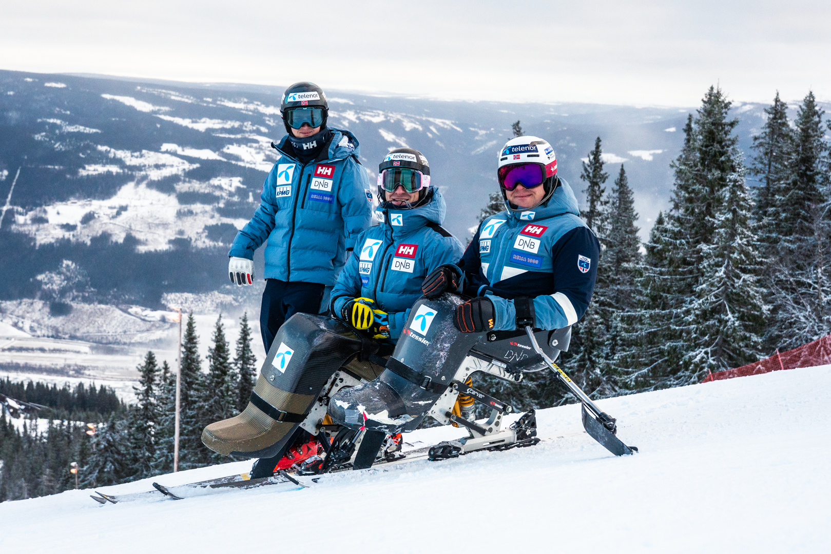 Tre parautøvere i alpinbakken med sitski og skiutstyr, omgitt av snødekt fjellandskap og grantrær.