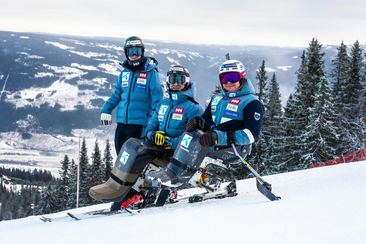 Tre parautøvere i alpinbakken med sitski og skiutstyr, omgitt av snødekt fjellandskap og grantrær.